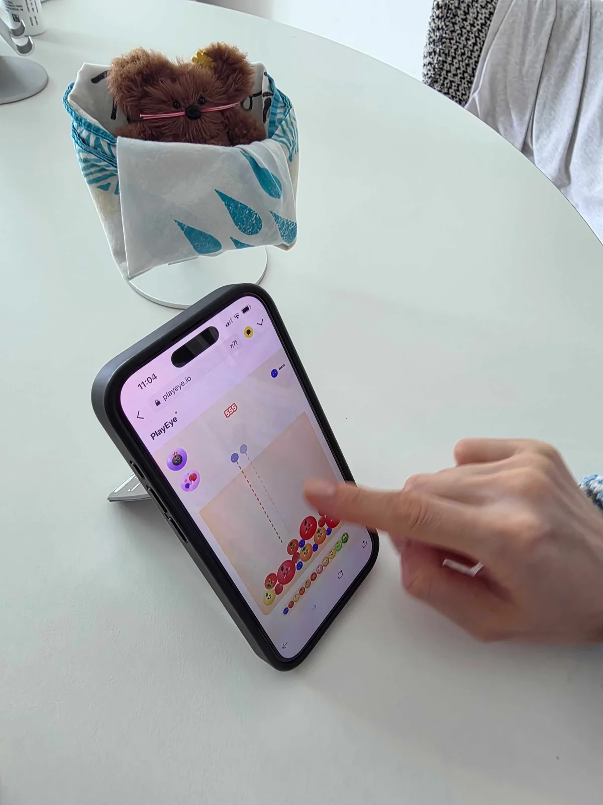 An iPhone propped on a kickstand on an office table showing Watermelon Game at 555 points, with a finger reaching toward the screen