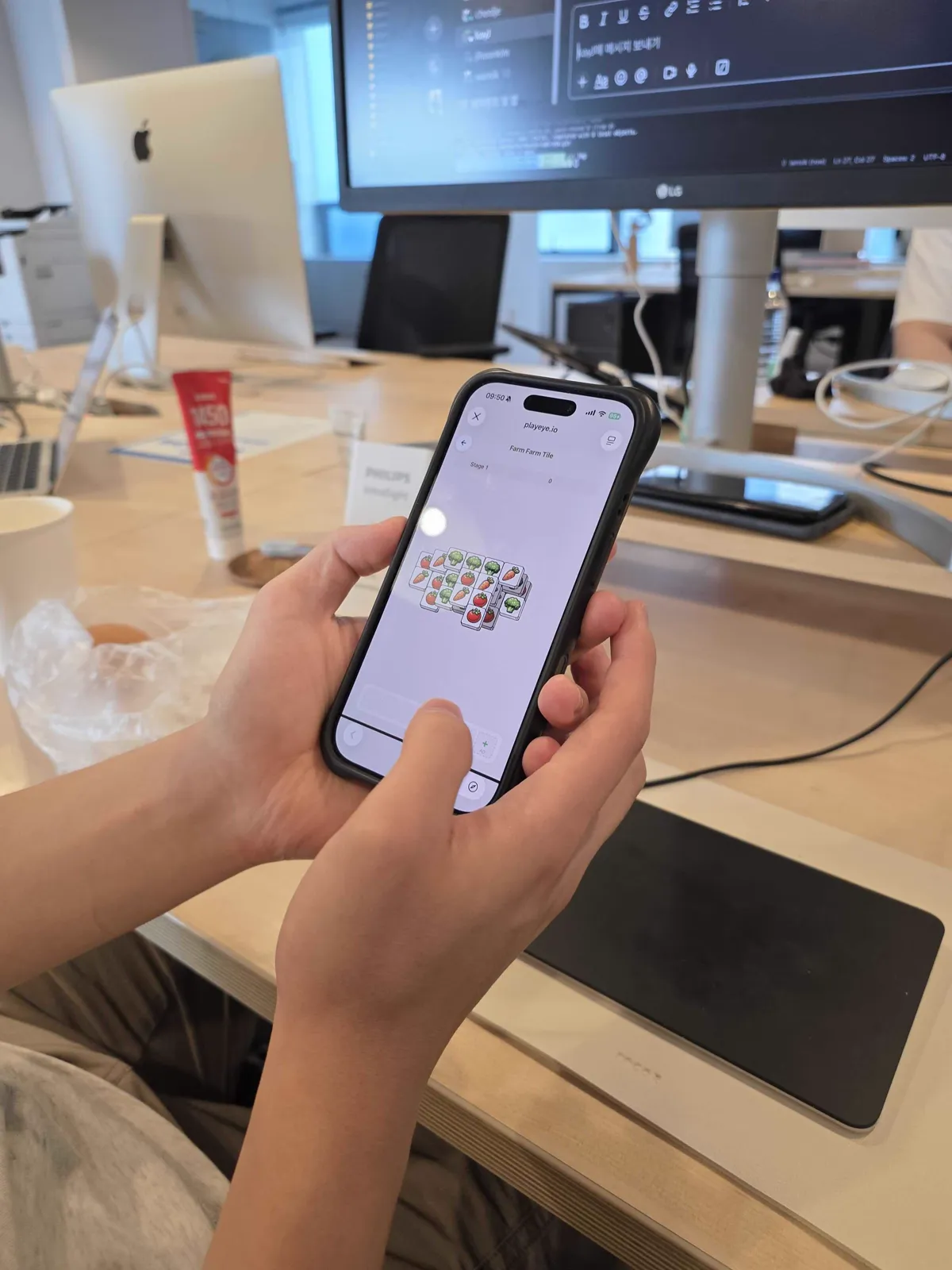 A player holding an iPhone with Farm Farm Tile Stage 1 open on the live PlayEye website in an office